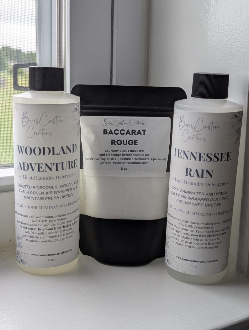 Op de afbeelding: Twee doorzichtige plastic flessen met zwarte doppen gevuld met vloeibaar wasmiddel met de labels Woodland Adventure en Tennessee Rain, naast een zwarte zak met Baccarat Rouge wasparfum, elk voorzien van witte etiketten met zwarte tekst.