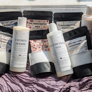 Op de afbeelding: Een collectie wasverzorgingsproducten waaronder twee witte plastic flessen vloeibaar wasmiddel met de labels Favorite Hoodie en The Narcissistic Man, naast diverse zwarte zakjes met wasparfum met de labels Favorite Hoodie, The Narcissistic Man en Cocobutter Cashmere.