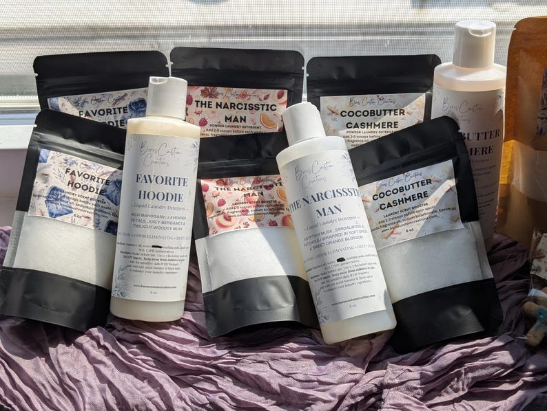 Op de afbeelding: Een collectie wasverzorgingsproducten waaronder twee witte plastic flessen vloeibaar wasmiddel met de labels Favorite Hoodie en The Narcissistic Man, naast diverse zwarte zakjes met wasparfum met de labels Favorite Hoodie, The Narcissistic Man en Cocobutter Cashmere.