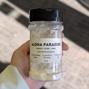 Può includere: Un flacone dosatore in plastica trasparente contenente polvere bianca, etichettato Aloha Paradise Odor Eliminating Dust. L'etichetta riporta profumi di ananas, cocco e mango, istruzioni per agitare, lasciare agire per 15+ minuti e aspirare, peso 170g.