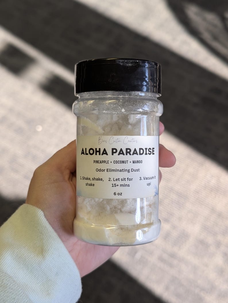 May include: A clear plastic shaker bottle filled with white powder labeled Aloha Paradise Odor Eliminating Dust. The label lists pineapple, coconut, and mango scents, instructions to shake, let sit for 15+ mins, and vacuum it up, with a net weight of 6 oz.