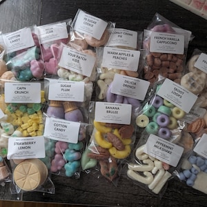 May include: A collection of wax melts in clear plastic bags, each labeled with a unique scent name. The melts are in various shapes and colors, including pink, blue, yellow, and brown. Scents include "Cotton Candy," "Strawberry Lemon," and "Blueberry Pancake."