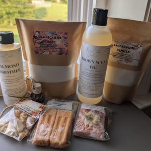 Może przedstawiać: Kolekcja produktów do prania, w tym dwie wysokie, przezroczyste plastikowe butelki z etykietami Almond Brother i Brown Sugar Fig, dwie brązowe papierowe torebki z etykietami Jamaican Me Crazy i Sandalwood Vanilla oraz trzy małe przedmioty owinięte w folię.