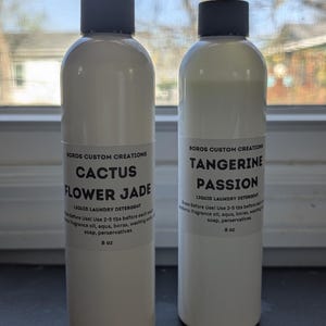 Op de afbeelding: Twee 8 oz witte plastic flessen vloeibaar wasmiddel met zwarte schroefdoppen. De etiketten vermelden Boros Custom Creations, waarbij de ene fles is gelabeld als Cactus Flower Jade en de andere als Tangerine Passion, inclusief gebruiksinstructies.
