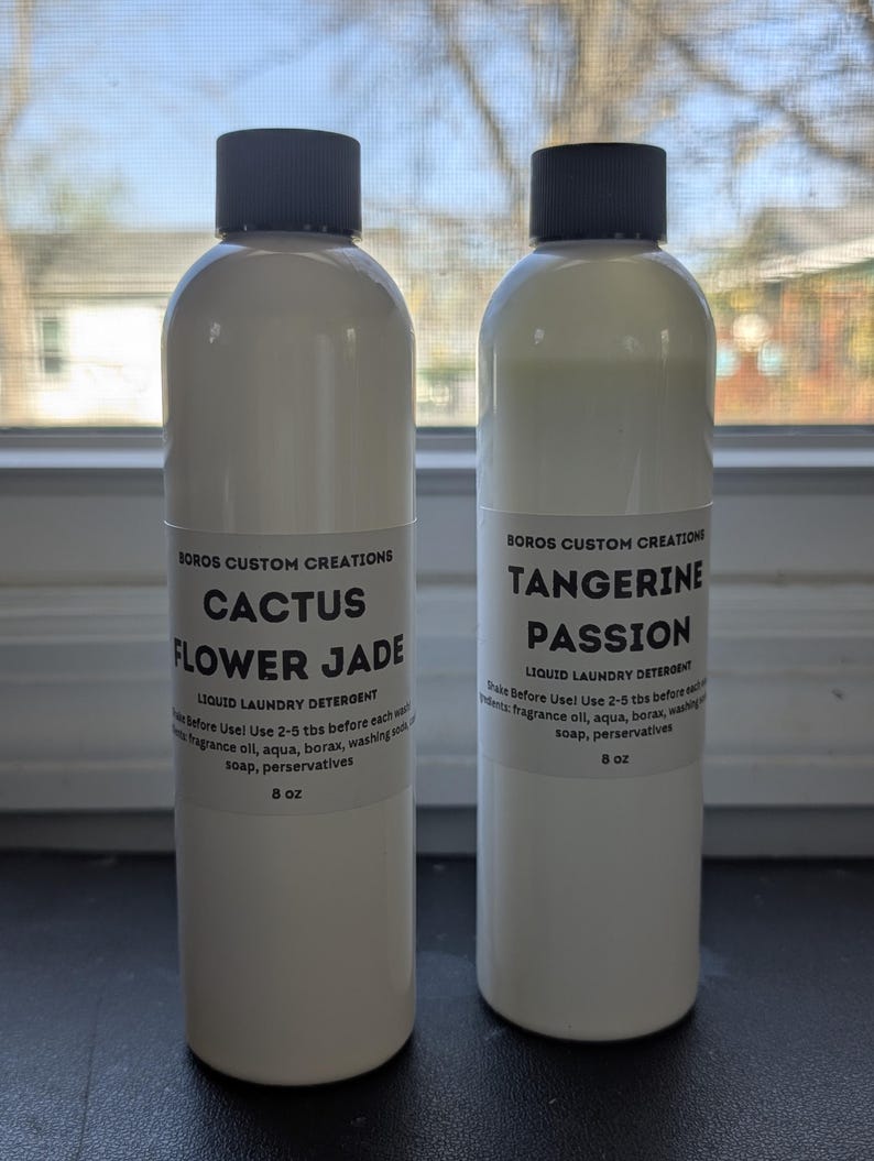 Op de afbeelding: Twee 8 oz witte plastic flessen vloeibaar wasmiddel met zwarte schroefdoppen. De etiketten vermelden Boros Custom Creations, waarbij de ene fles is gelabeld als Cactus Flower Jade en de andere als Tangerine Passion, inclusief gebruiksinstructies.