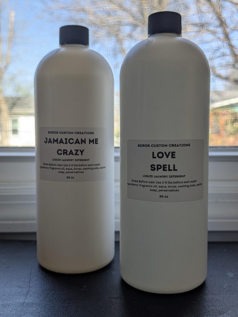 Op de afbeelding: Twee witte plastic flessen vloeibaar wasmiddel met zwarte doppen. Elke fles heeft een etiket met de tekst Boros Custom Creations, de ene gelabeld als Jamaican Me Crazy en de andere als Love Spell, beide met een inhoud van 1065 ml.