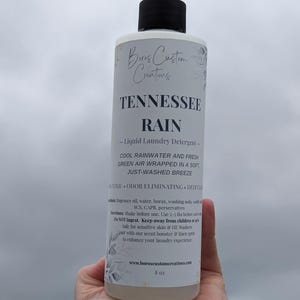 Op de afbeelding: Een fles van 8 oz Boros Custom Creations Tennessee Rain vloeibaar wasmiddel met een zwarte dop. Het witte etiket bevat zwarte tekst die de geur beschrijft als koel regenwater en frisse groene lucht, samen met instructies voor gebruik.