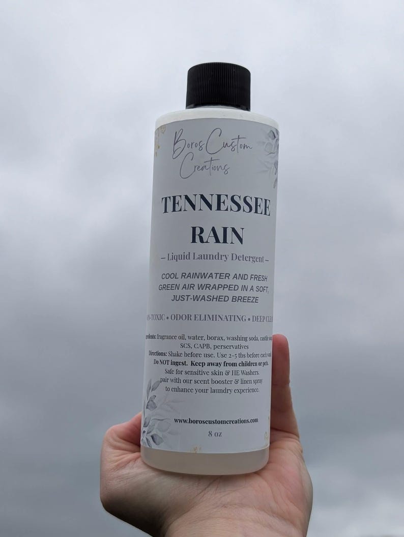 Op de afbeelding: Een fles van 8 oz Boros Custom Creations Tennessee Rain vloeibaar wasmiddel met een zwarte dop. Het witte etiket bevat zwarte tekst die de geur beschrijft als koel regenwater en frisse groene lucht, samen met instructies voor gebruik.