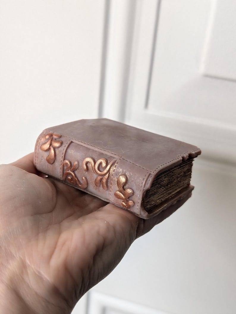 May include: A small, decorative book made of faux leather with a distressed, vintage look. The book is a light brown color with gold accents and intricate designs on the cover. Book Wax Melt Bookish Decor Book Lover Gift Bookish Wax Melt Cozy Gift for Teacher Booktok Fantasy Lovers Literary Decor Gifts for Readers