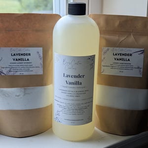 Op de afbeelding: Een set van drie wasproducten, waaronder twee bruine papieren zakjes gevuld met wit poeder en een doorzichtige plastic fles met lichtgele vloeistof. Alle labels bevatten de tekst Lavender Vanilla en de merknaam Boss Custom Creations.