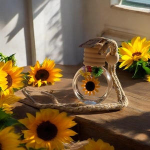 May include: A clear glass car air freshener with a sunflower and other flowers inside. The air freshener is hanging from a brown rope with a wooden bead. The air freshener is surrounded by yellow sunflowers.