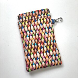 Peut inclure: Une petite pochette avec une fermeture à glissière et un imprimé de crayons colorés. La pochette a un anneau en métal argenté et une sangle bleue avec un crochet en métal argenté.