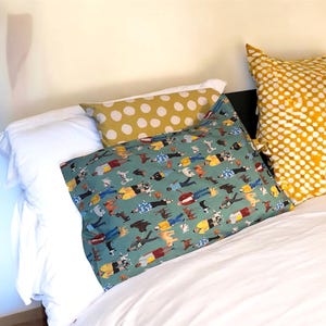 Puede incluir: Una cama con sábanas blancas y varias almohadas. Una almohada tiene un fondo verde azulado con un patrón de perros y personas. Otras dos almohadas tienen un fondo amarillo con lunares blancos.