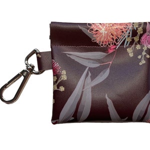 May include: A small brown pouch with a floral print featuring pink and white flowers and leaves. The pouch has a silver clip attached to it.