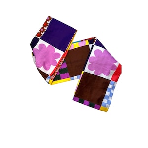 Puede incluir: Una bufanda colorida con un diseño de patchwork. Presenta cuadrados de color púrpura, marrón y blanco, adornados con patrones florales y geométricos. La bufanda está hecha de un material suave, adecuada para diversas ocasiones.