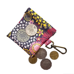 Könnte beinhalten: Eine kleine, schwarze Tasche mit einem bunten Blumenmuster und einem goldfarbenen Reißverschluss. Die Tasche hat einen goldfarbenen Metallclip. Um die Tasche herum liegen mehrere australische Münzen.