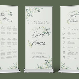 May include: Three white wedding signage boards with green floral designs and gold accents. The first board has a seating chart with table numbers and guest names. The second board has the text "Welcome to the Wedding of Gary and Emma" with the date "10th October 2020". The third board has the text "What Where" and a list of wedding events with times.