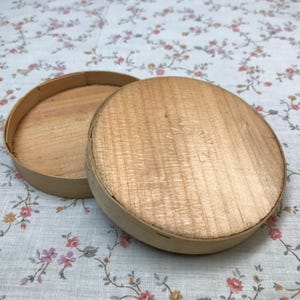 May include: An open, round, wooden box with a lid. The box and lid are made of light-colored wood. The box is sitting on a floral patterned fabric. The lid is slightly ajar, revealing the interior of the box.