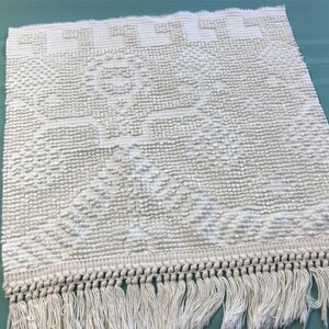 May include: A square off-white textile mat featuring a raised tufted texture with a central geometric tree-like motif and a Greek key border pattern. The bottom edge is finished with a row of thick, knotted fringe tassels hanging from the base.
