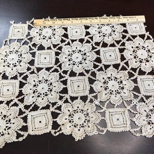 May include: An off-white crocheted doily featuring a repeating pattern of circular floral motifs and small squares, measuring approximately 12 inches in width as indicated by the wooden ruler placed along the top edge of the textile piece.