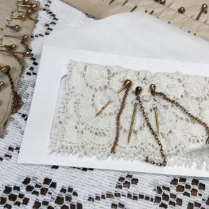 May include: An assortment of antique hat pins and chain pins, some with decorative ball and chain accents, displayed on a white lace doily inside a white envelope. Additional pins are arranged on a piece of aged paper.