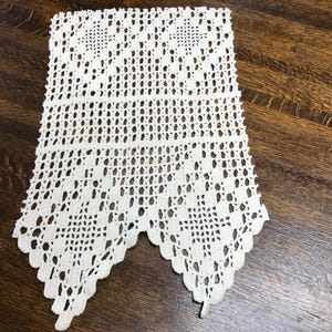 May include: Cream-colored, handmade crochet doily with a detailed openwork design. The rectangular piece features a scalloped edge and intricate patterns, showcasing the delicate craftsmanship. The doily is placed on a dark wooden surface.