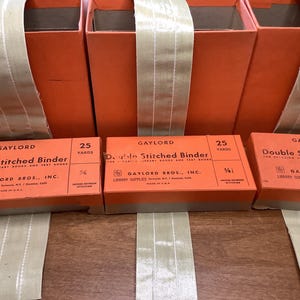 May include: Three orange boxes of Gaylord Double Stitched Binder with beige ribbon. Each box has the text "Double Stitched Binder" and "25 Yards". The boxes are for library books and text books.