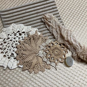 May include: An assortment of handmade crochet doilies in cream and tan colors, along with a length of beige lace trim, a small oval button, and a piece of corrugated cardboard. The doilies feature intricate floral and geometric patterns.