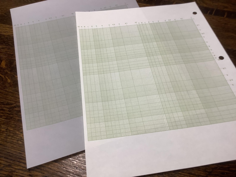 May include: Two sheets of graph paper with a grid pattern. One sheet is a light gray color, and the other is a light green color. The sheets are on a wooden surface. The paper has holes along the left edge.