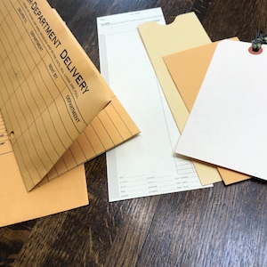 May include: An assortment of stationery items, including manila envelopes with the text "R-DEPARTMENT DELIVERY", blank tags, and various paper sheets. The items are arranged on a dark wooden surface.