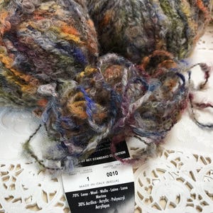 May include: Close-up of colorful yarn balls with a mix of orange, blue, green, and purple hues. The yarn has a textured, slightly fuzzy appearance. A label with text is visible, indicating the yarn's composition and origin.