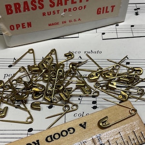 May include: A collection of brass safety pins scattered on sheet music, with a wooden ruler and a box labeled "BRASS SAFETY PINS." The box also reads "RUST PROOF" and "GILT" and "MADE IN U.S.A."