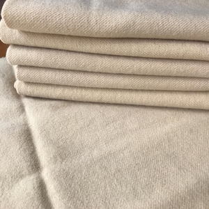 May include: A stack of five folded pieces of cream-colored fabric with a visible diagonal twill weave pattern. The fabric has a soft, slightly fuzzy texture and is arranged neatly on top of a larger, unfolded piece of the same matching material.