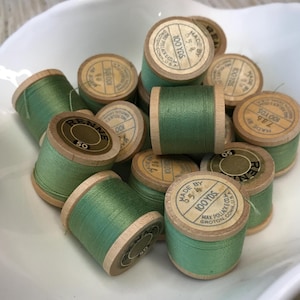 May include: A collection of vintage wooden spools wrapped in sage green thread. Each spool features a circular paper label on the end with printed text including Made By Max Pollack & Co. Groton, Conn. U.S.A. and 100 Yds, or the brand Renne.