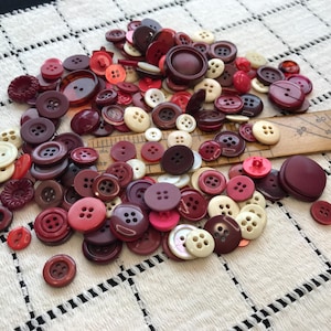 May include: A large collection of assorted round buttons in shades of red, burgundy, maroon, and cream. The buttons vary in size, texture, and hole count, with some featuring four holes and others two. A wooden ruler is visible, showing measurements.