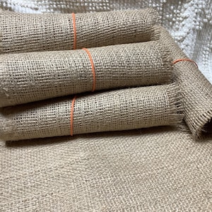 May include: Several rolls of natural burlap fabric, secured with orange rubber bands. The burlap is a light brown color and has a coarse, woven texture. The rolls are stacked on top of a larger piece of the same fabric.