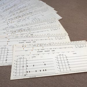 May include: A stack of vintage student request cards with printed text and grid patterns. The cards are off-white with black ink, featuring fields for codes, courses, and student information. The cards are arranged on a neutral-toned surface.