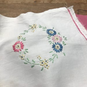 May include: A square white fabric piece featuring a central embroidered floral wreath. The wreath consists of small pink, blue, and yellow flowers with green stitched leaves and stems. The fabric has raw, unfinished edges and is layered over pink fabric.
