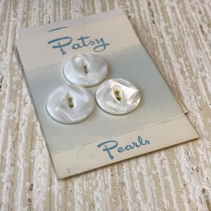 May include: Three round, white, iridescent buttons with a geometric, faceted surface design attached to a rectangular card. The card features the blue printed text "Patsy" at the top and "Pearls" at the bottom, with a light blue gradient background.