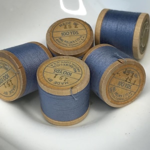 May include: Five wooden spools of blue sewing thread, each featuring a circular paper label printed with 'MADE BY MAX POLLACK & CO. GROTON, CONN., U.S.A.' and '100 YDS.' The thread is wound tightly around the cylindrical wooden cores.