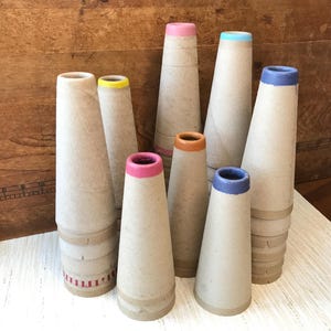 May include: A collection of eight conical cardboard yarn spools arranged in a cluster. Each spool features a light tan paper texture with a distinct colored rim, including shades of pink, yellow, orange, blue, and purple, with one base showing red stripes.