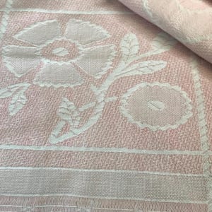 May include: A light pink and white woven fabric featuring a repeating grid pattern. Each square contains a raised, textured floral motif with a large five-petaled flower and a smaller circular bloom, all rendered in a soft, monochromatic color palette.