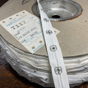 May include: A spool of white fabric trim with brass studs. The label reads "Yale T117 White 1 1/0 Brass Stud 144 YDS. DOZ. 2F-83 WINDO".