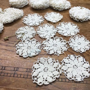 May include: A collection of small, round, off-white crocheted lace doilies featuring a floral pattern, arranged alongside a gold-toned safety pin. A wooden ruler is visible at the bottom, showing measurements in inches for scale.
