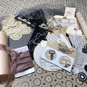 May include: A collection of craft supplies including a rolled parchment paper, a dusty rose silk ribbon, cream-colored crochet lace, black lace trim, vintage music sheets, metal paper clips, a small key in a coin holder, and assorted buttons.