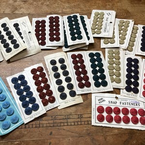 May include: A collection of vintage silk-covered snap fasteners on paper cards in various colors including red, blue, black, and beige. Each card features the text "Non-Ravel Snap Fasteners" and "Made in Korea" or "Made in Japan".