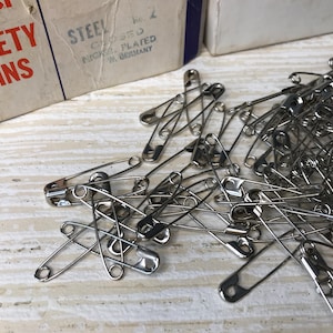 May include: A collection of silver-colored safety pins scattered on a light-colored surface. Two vintage cardboard boxes are visible, one labeled "SAFETY PINS" in red letters. The boxes have blue trim and text that reads "STEEL CLOSED NICKEL PLATED W. GERMANY."