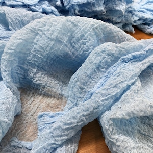 May include: A light blue fabric runner with a crinkled, textured surface. The material is semi-sheer and lightweight, featuring a loose, open weave pattern that creates a series of folds and ripples across the length of the textile.