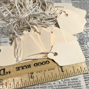 May include: A collection of blank, cream-colored tags with string, resting on a vintage dictionary page. A wooden ruler with inch markings is positioned below the tags, with the text "GOOD RULE" visible.