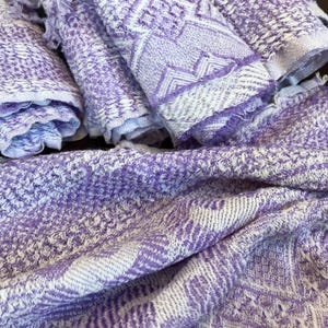 May include: A collection of woven textiles featuring a light purple and white geometric pattern. The fabric displays a textured, intricate weave with visible fringe edges on the rolled pieces, showcasing a repeating diamond and chevron design.